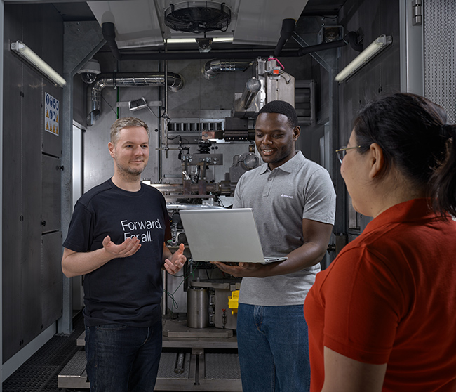 Three people standing in a factory having a conversation and one person holding a laptop